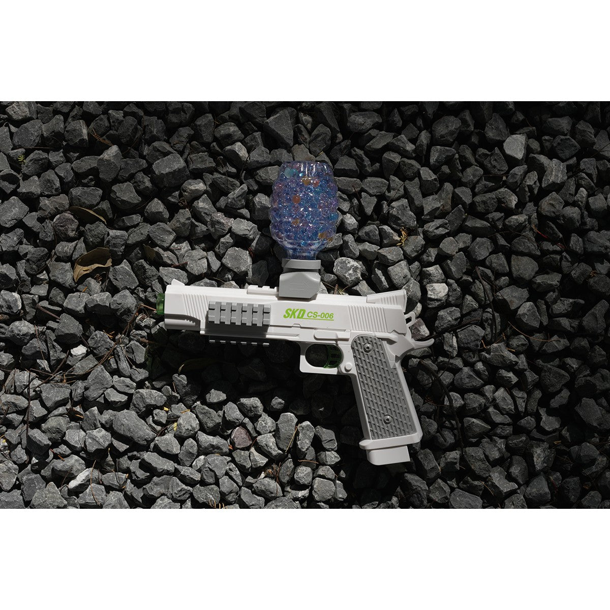 SKD CS006S M1911 Electric Gel Blaster placed on gravel with blue gel beads in hopper, suitable for outdoor toy combat scenarios.